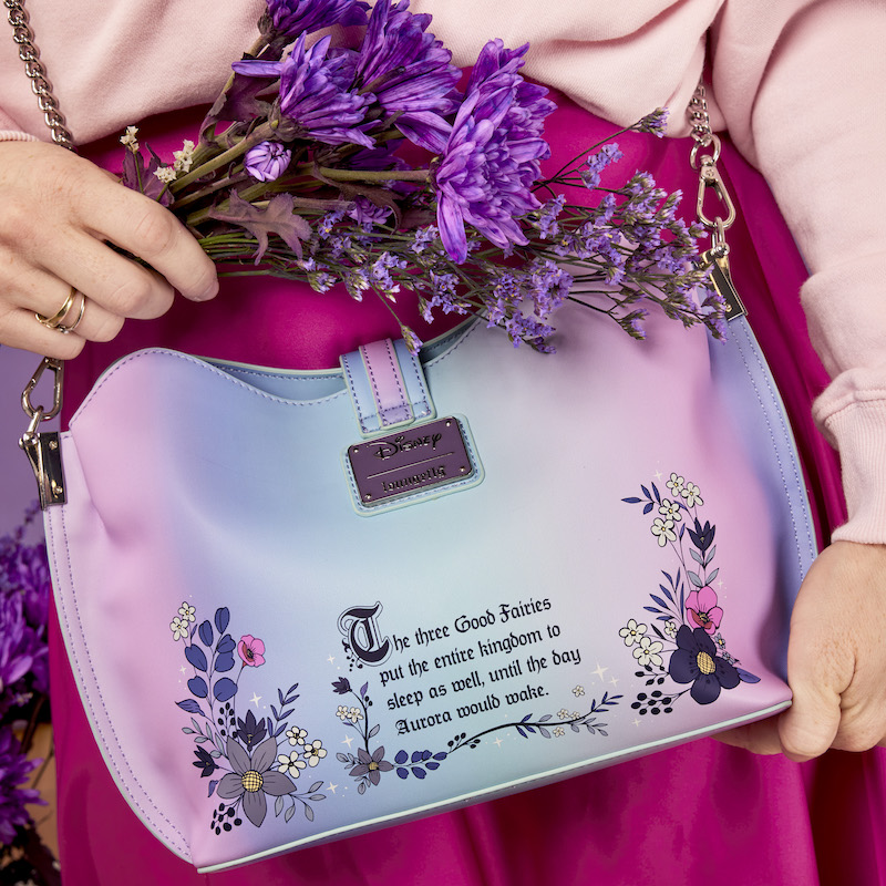 Image of someone wearing a pink sweater and skirt holding flowers and wearing the Loungefly Disney Sleeping Beauty 65th Anniversary Floral Ombre Crossbody Bag, showing off the back of the bag with a quote from Sleeping Beauty embroidered on it. 